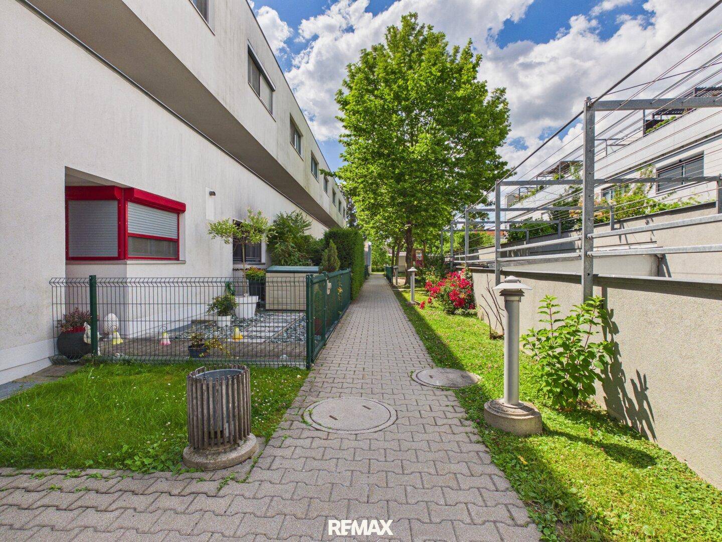 Gepflasterter Weg zwischen Gebäuden mit kleinen privaten Gartenbereichen und grünen Hecken.