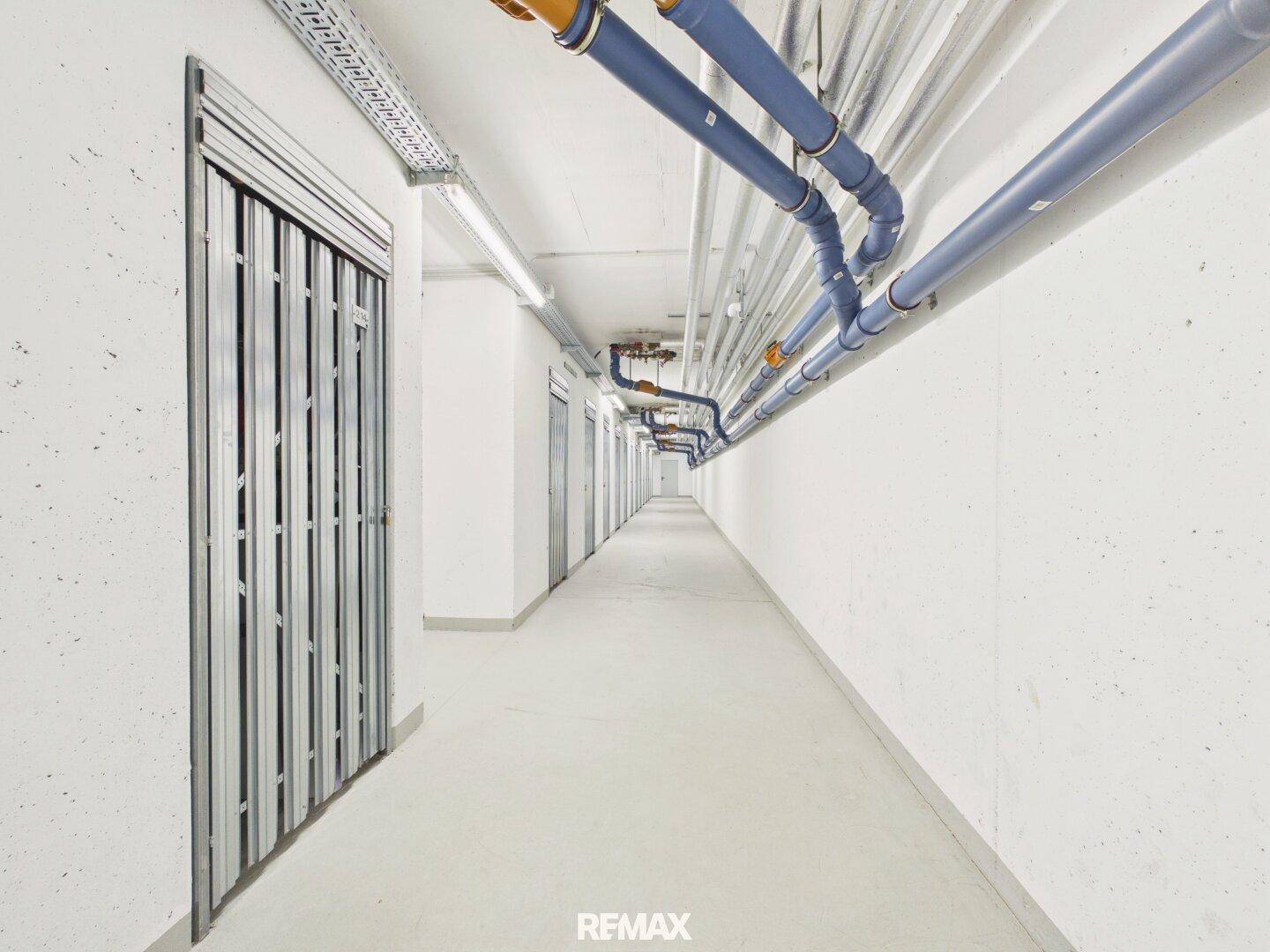Langer Kellerflur mit individuellen Abstellräumen und sichtbaren Versorgungsleitungen an der Decke.