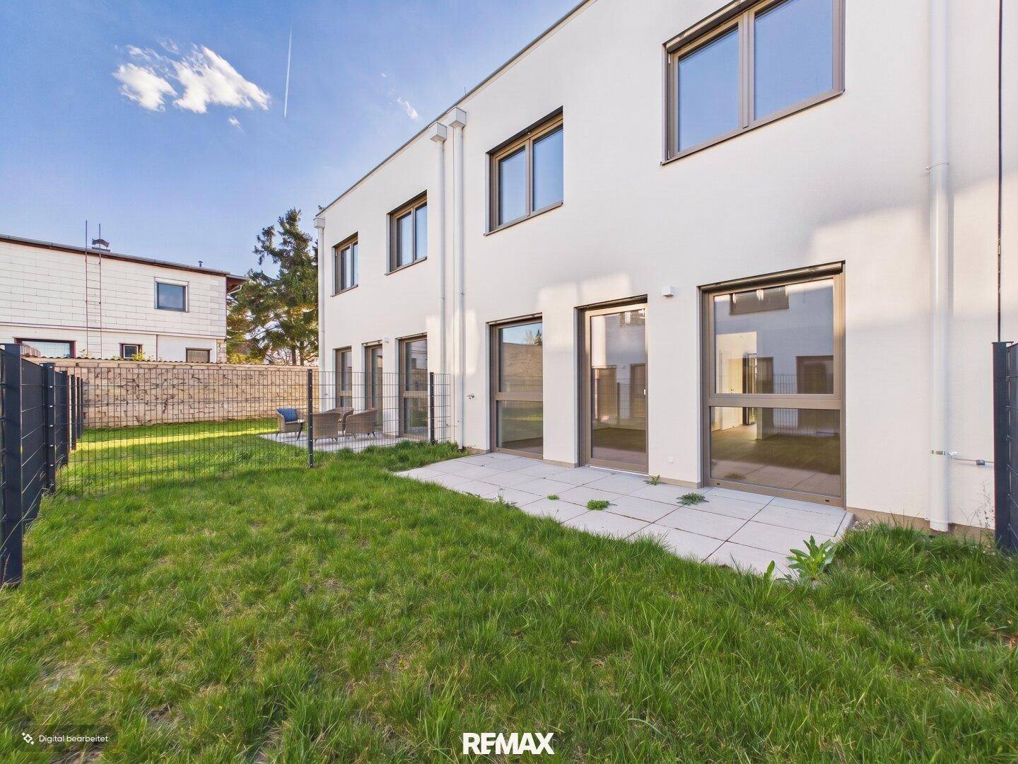 Rückseite eines modernen Gebäudes mit Terrasse und Rasenfläche, zugänglich durch große Fenstertüren.