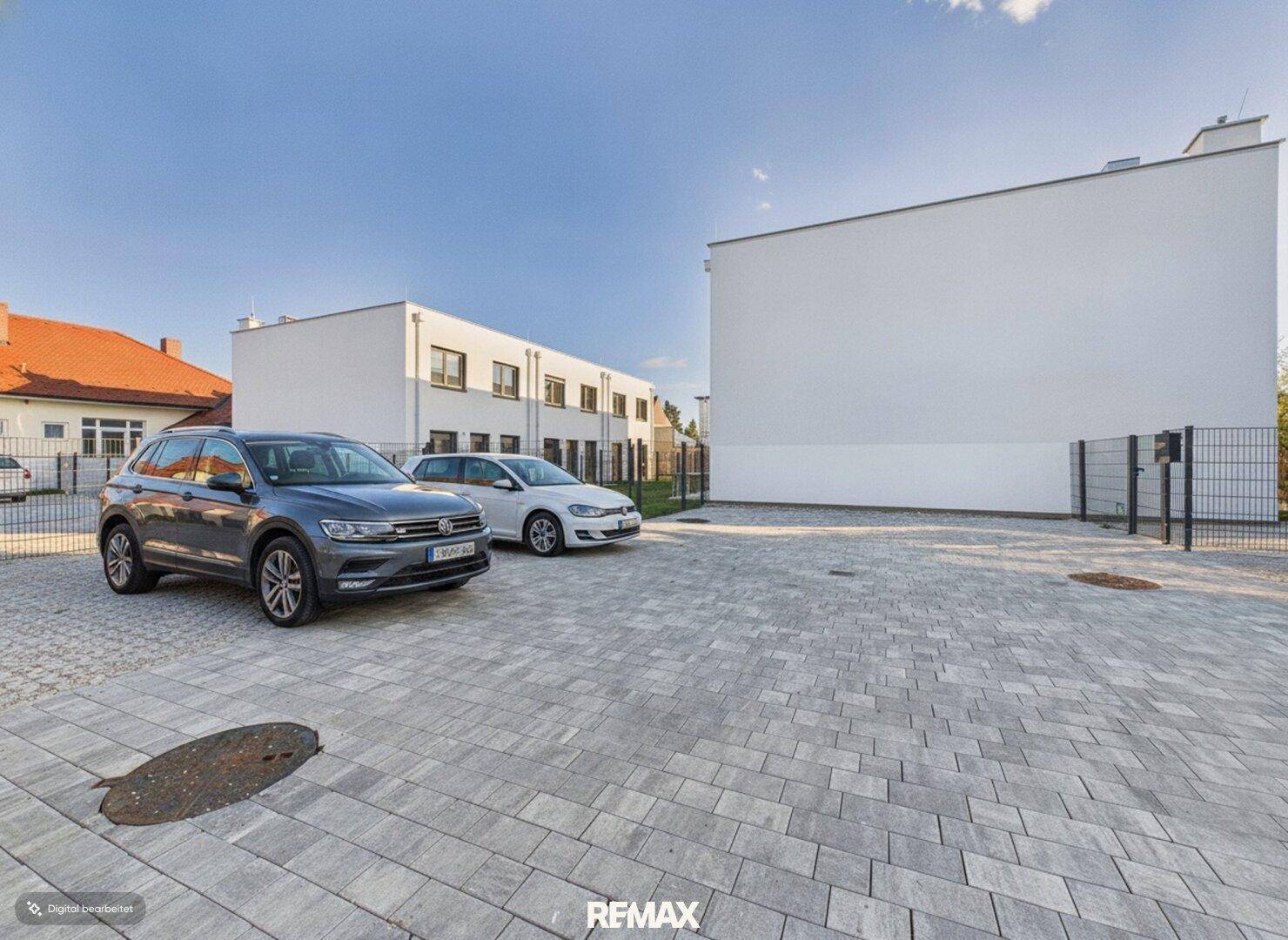Außenansicht mit gepflastertem Parkplatz und modernen weißen Gebäuden unter blauem Himmel.