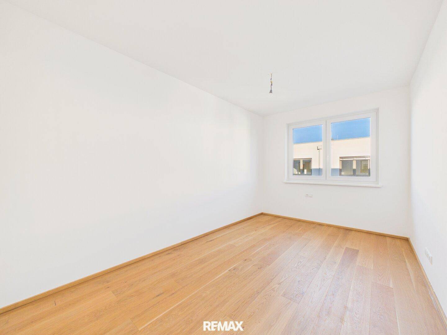 Leeres Zimmer mit Holzboden und einem Fenster, das den Blick auf benachbarte Gebäude freigibt.