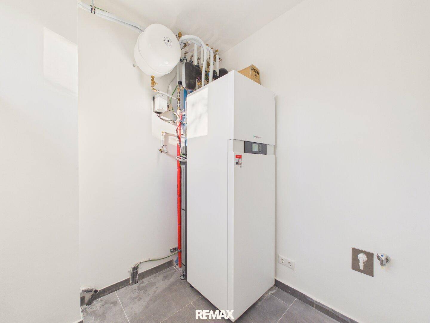 Technikraum mit moderner Heizungsanlage und Warmwasserspeicher auf gefliestem Boden.