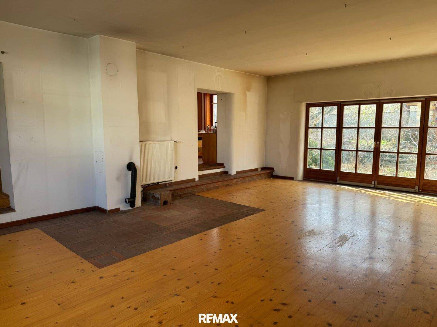 Heller Wohnraum mit Holzboden, Heizkörper und großen Fenstertüren zum Außenbereich.