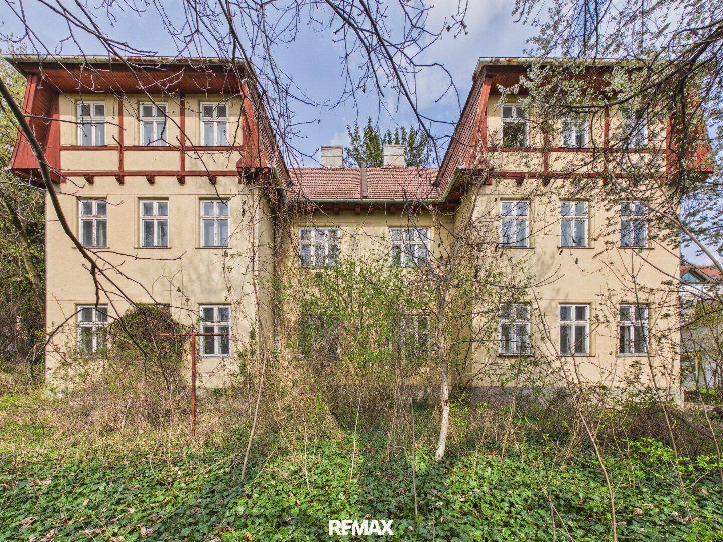 Rückansicht eines historischen Gebäudes mit Mansarddächern und verwildertem Gartenbereich.