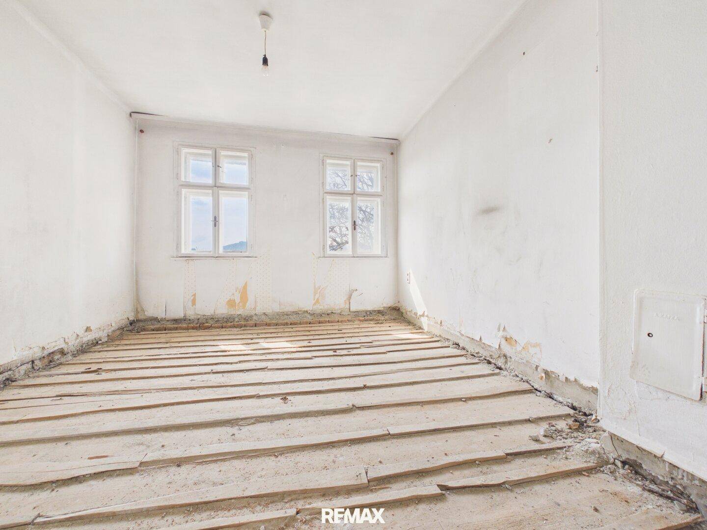 Entkernter Raum mit zwei Fenstern und freigelegten Holzdielen, wartet auf den Ausbau.
