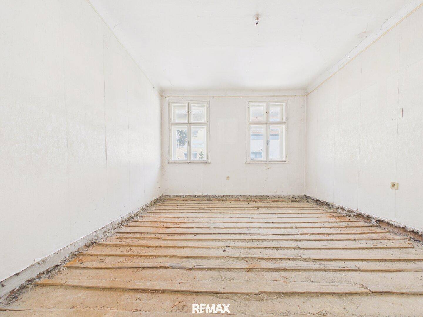 Großer Raum im Rohbau mit zwei Fenstern und freigelegten Holzdielen, wartet auf den Ausbau.