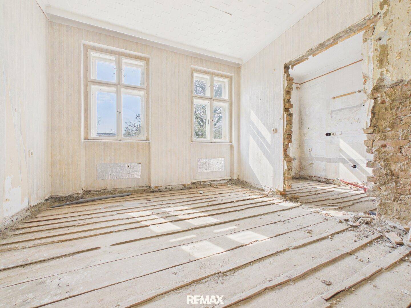 Großer Raum im Rohbau mit zwei Fenstern und freigelegten Holzdielen, bereit zur Sanierung.