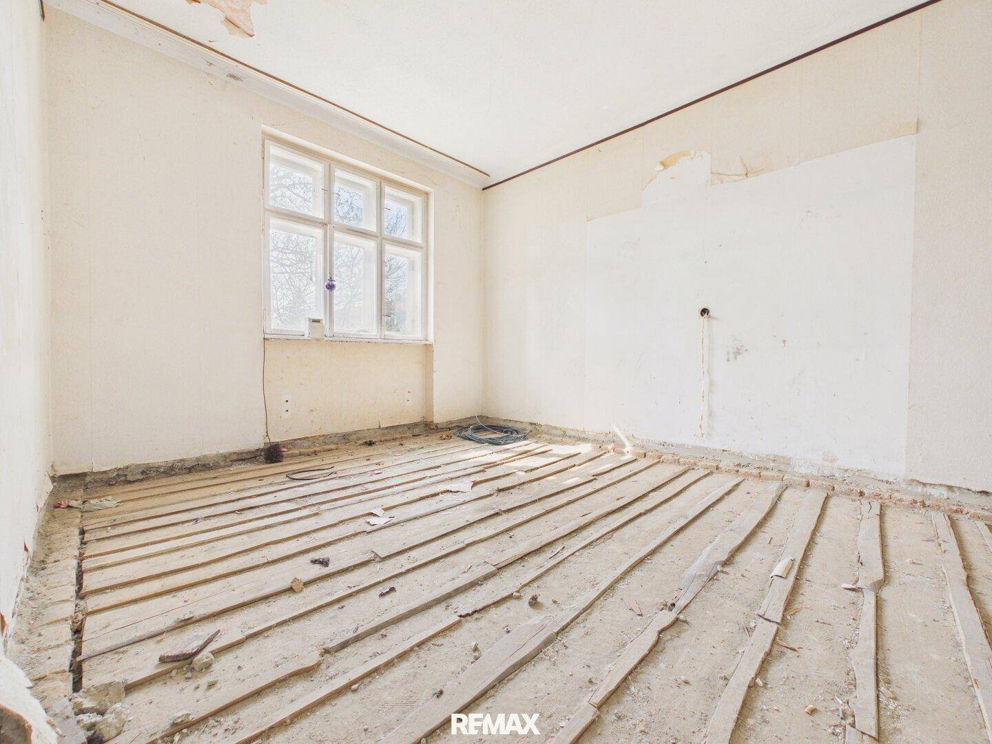 Raum im Rohbau mit einem Fenster und freigelegten Holzdielen, bereit für den Innenausbau.