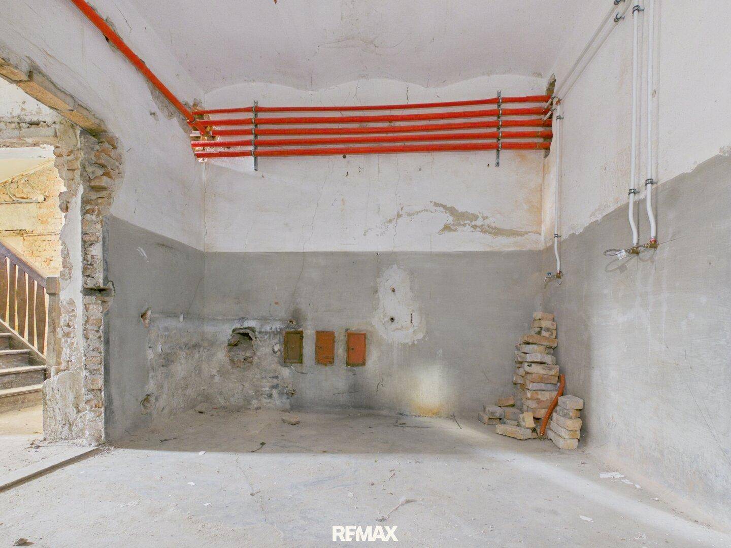 Kellerraum im Rohbau mit Betonboden, sichtbaren Heizungsrohren und Treppenaufgang.