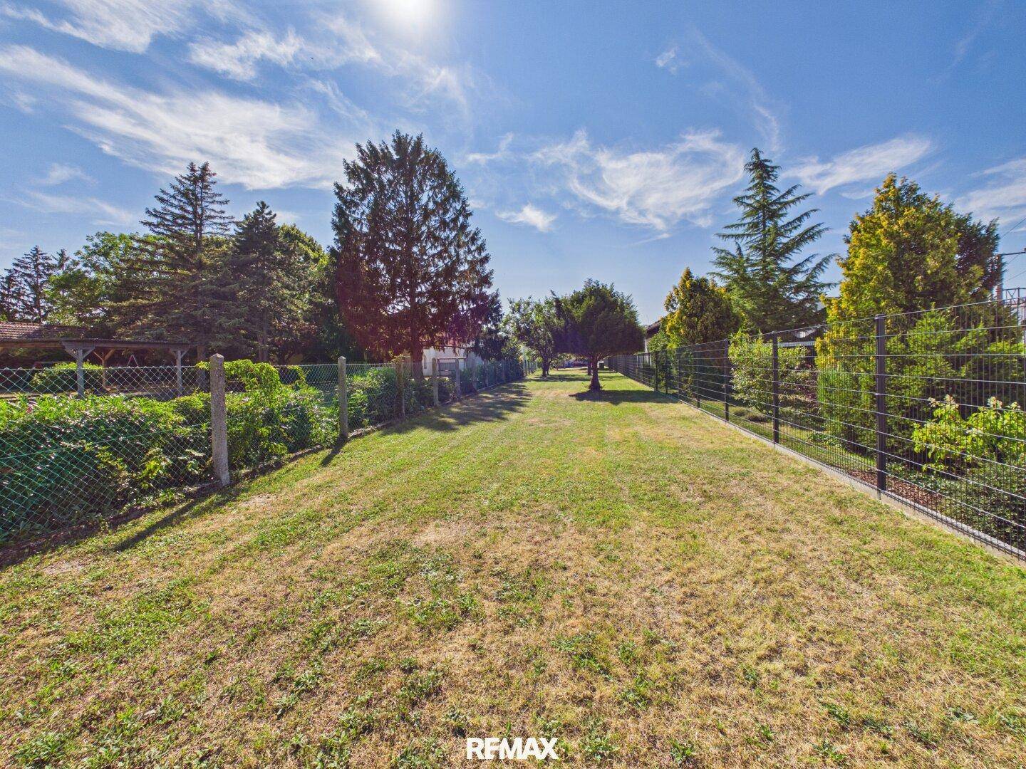 Langer, gepflegter Gartenbereich mit Rasen, Bäumen und Zäunen unter blauem Himmel.