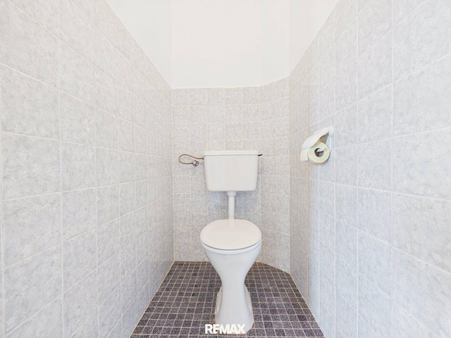 Kleines WC mit hellgrauen Fliesen an Wänden und dunklerem Fliesenboden.