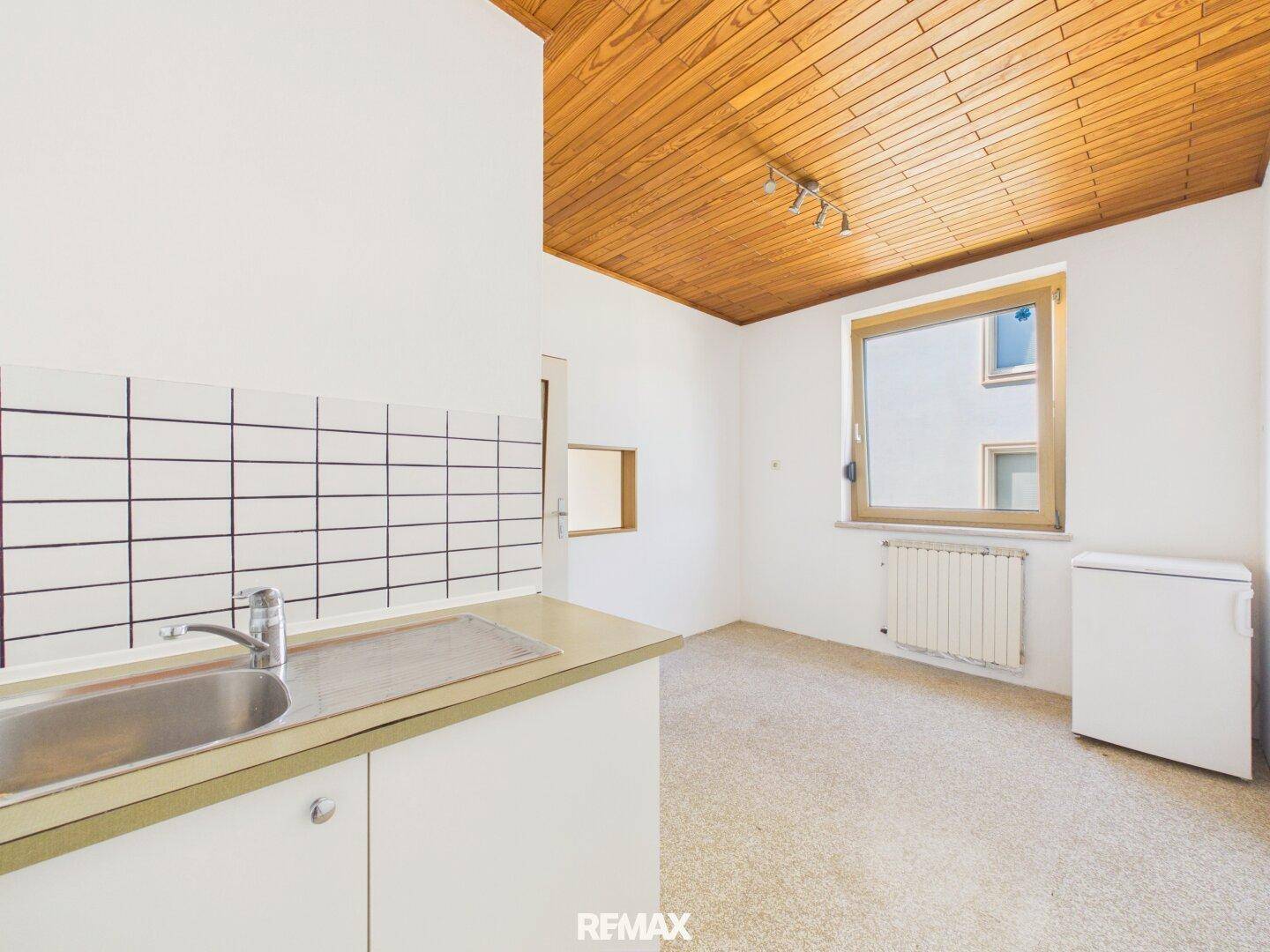 Küchenzeile mit Spüle, Fliesenrückwand und Holzpaneeldecke, hell durch Fenster.