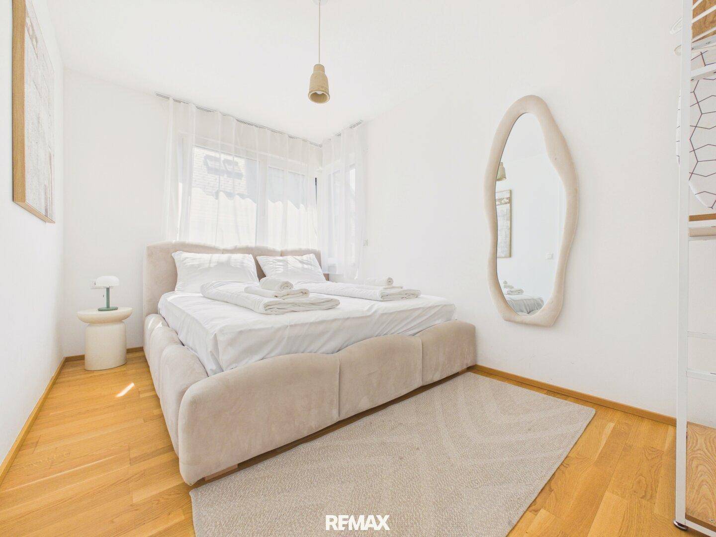 Gemütliches Schlafzimmer mit Doppelbett, Holzboden und einem großen, organisch geformten Spiegel.