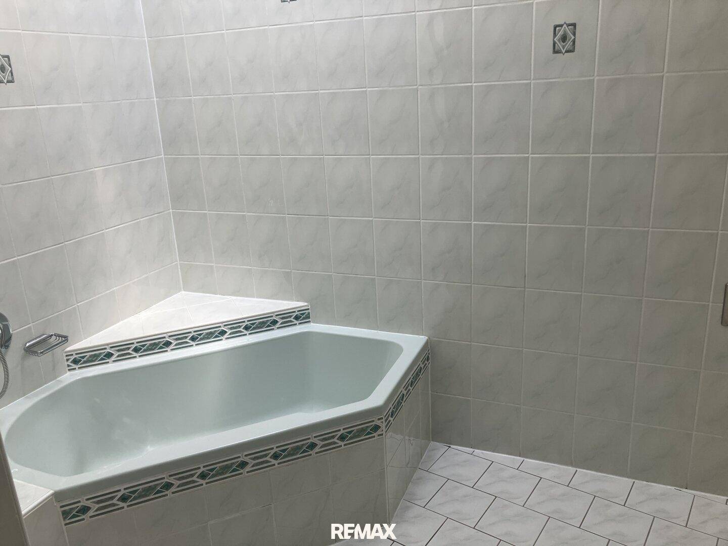 Badezimmer mit einer Eckbadewanne und hellen Fliesen an Wänden und Boden, im Stil der 80er Jahre.