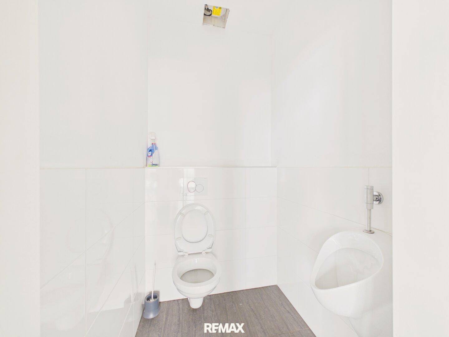 Toilettenraum mit wandhängendem WC und Urinal, ausgestattet mit weißen Fliesen und dunklem Boden.