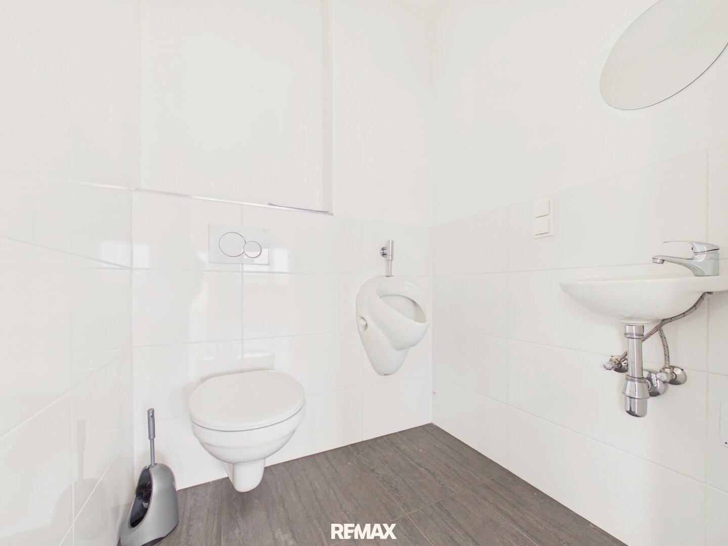 Helle Toilette mit Urinal, WC und Waschbecken, ausgestattet mit weißen Fliesen.