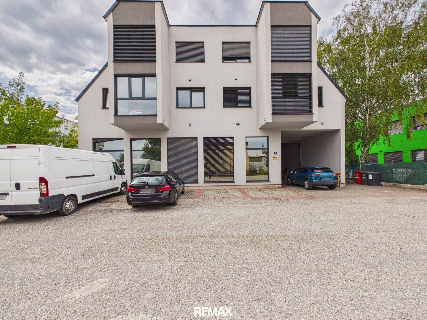 Modernes Geschäftsgebäude mit Parkplätzen und Zufahrt, umgeben von Bäumen.