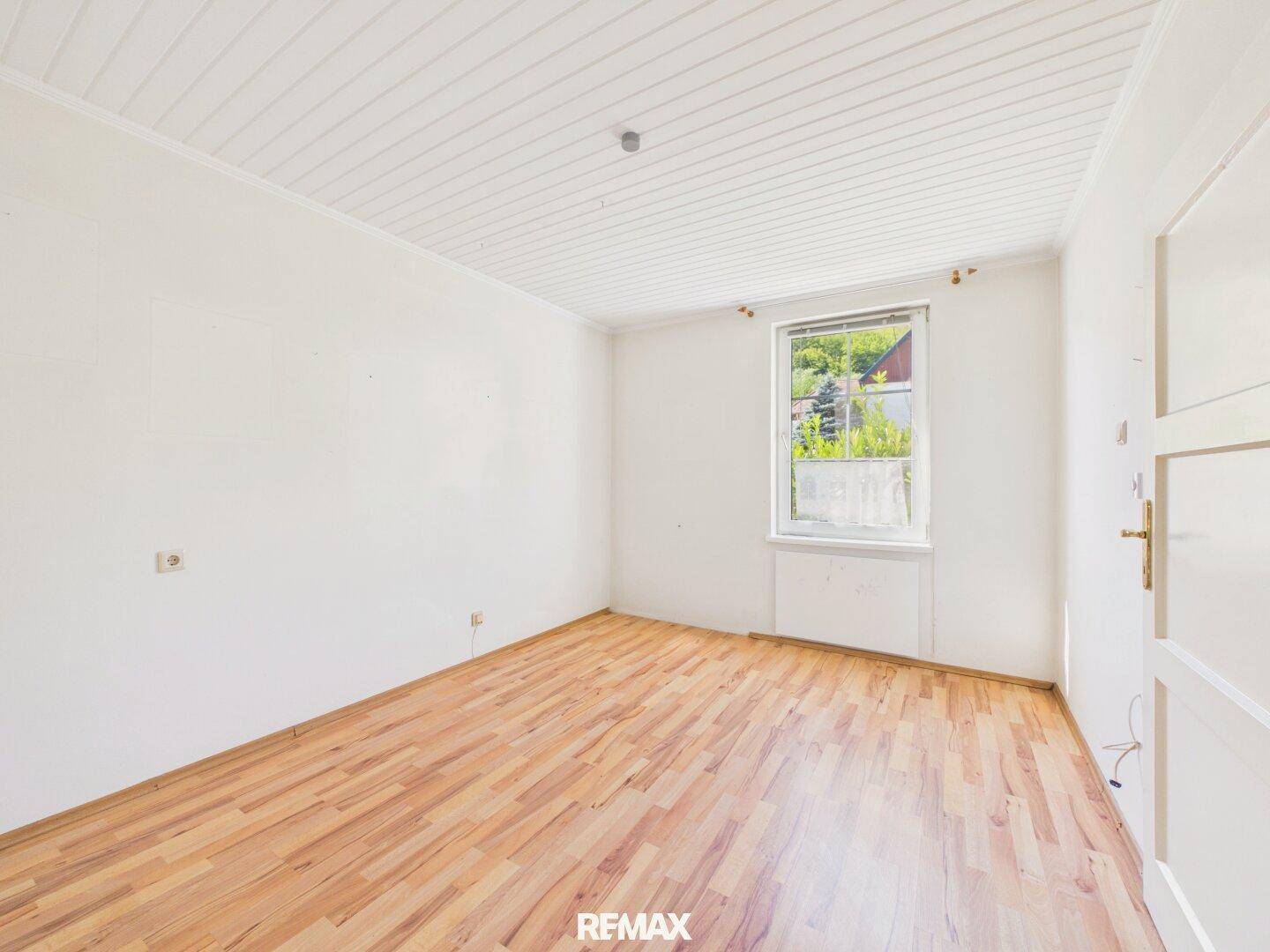 Einfaches Schlafzimmer mit Laminatboden und einem Fenster mit Blick ins Grüne.