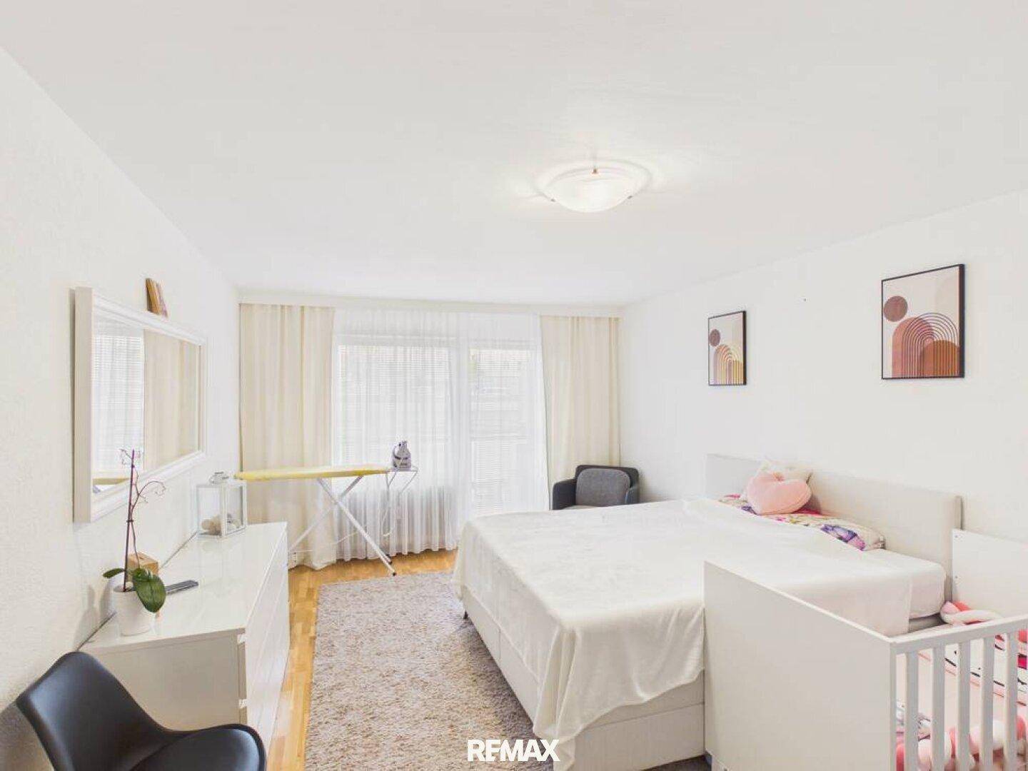 Helles Schlafzimmer mit großem Bett, Babybett und Fenster, ausgestattet mit einem Holzboden.
