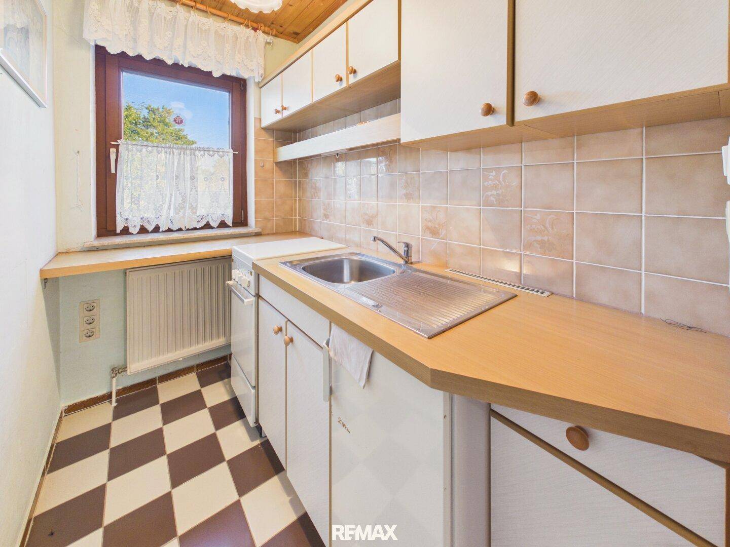 Kleine Küche mit kariertem Boden, Holztheke und Fenster mit Vorhängen.