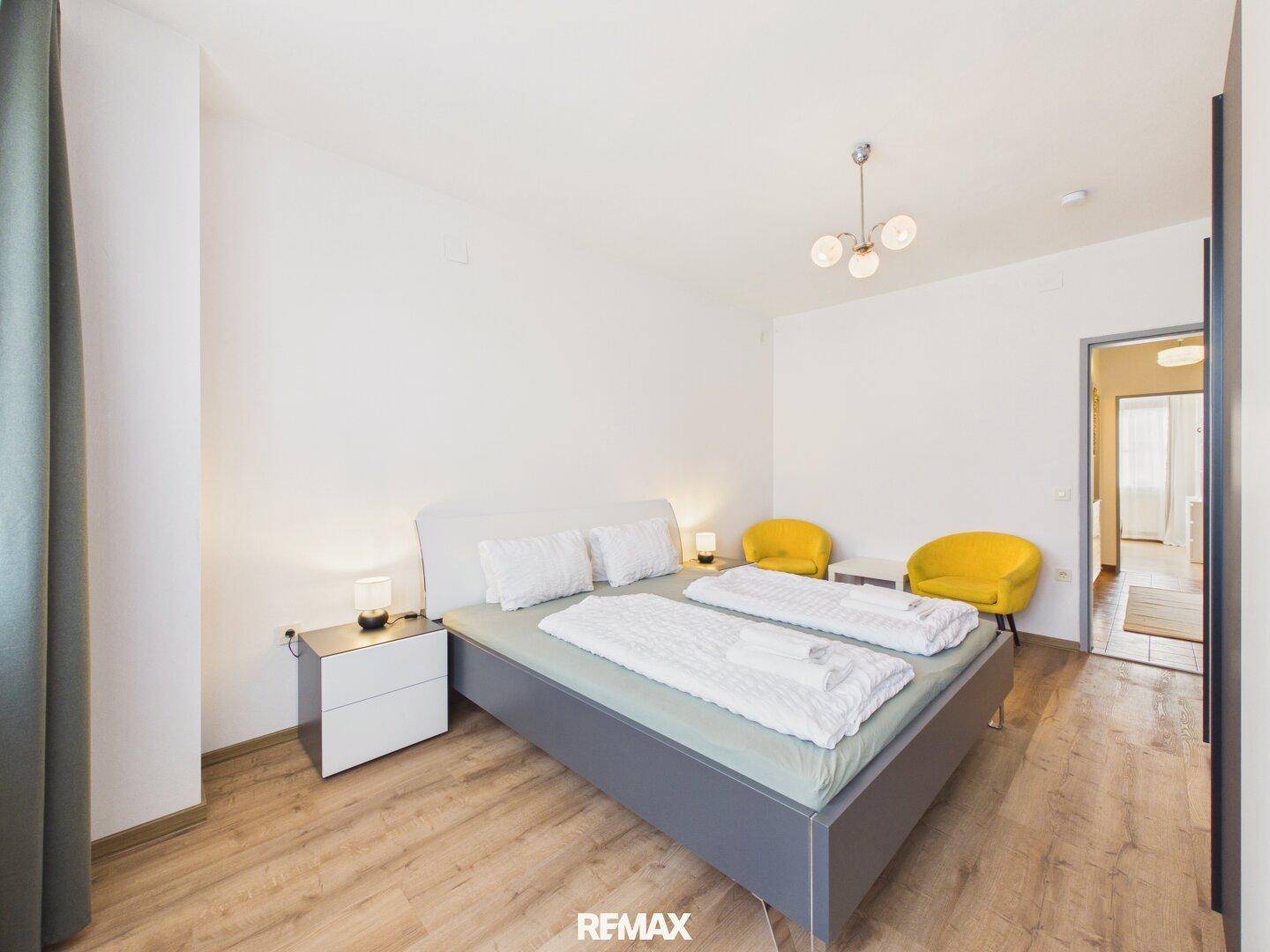 Modernes Schlafzimmer mit grauem Bett, Holzboden und zwei gelben Akzentstühlen.
