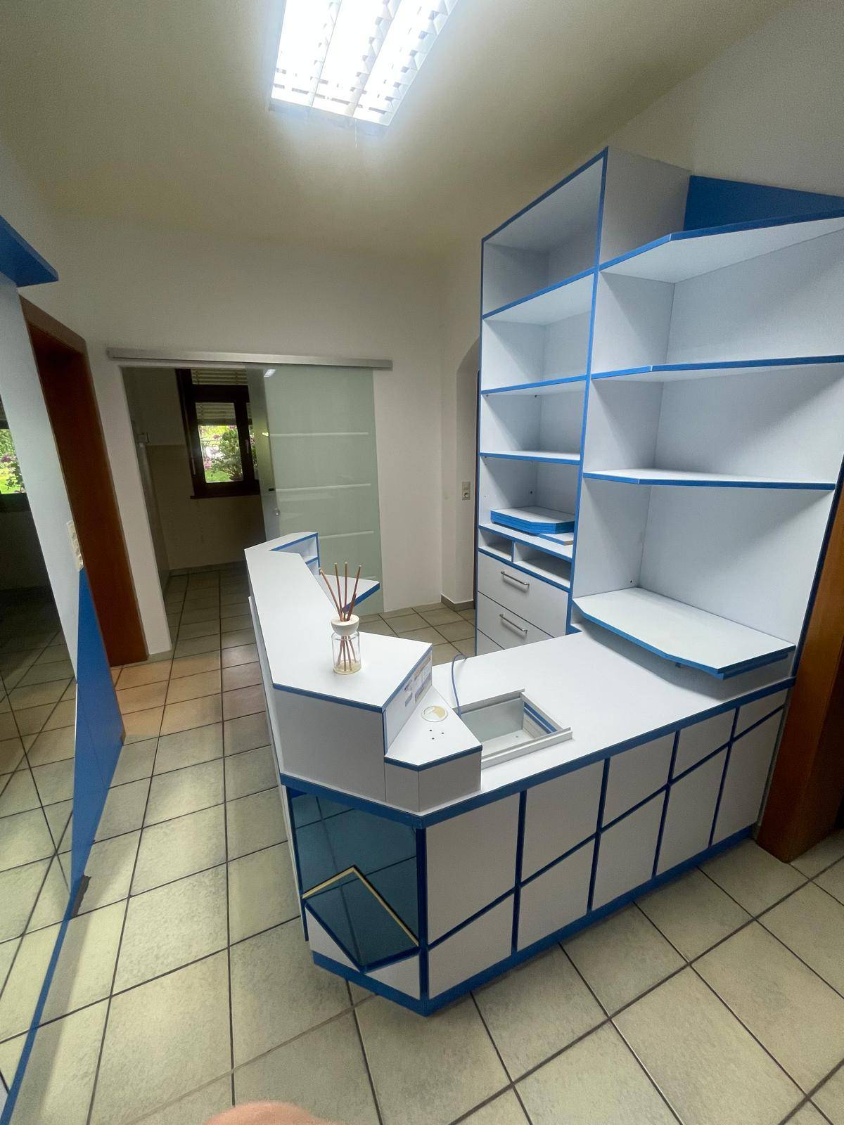 Empfangsbereich mit einer weiß-blauen Theke, Regalen und einer Glasschiebetür im Hintergrund.