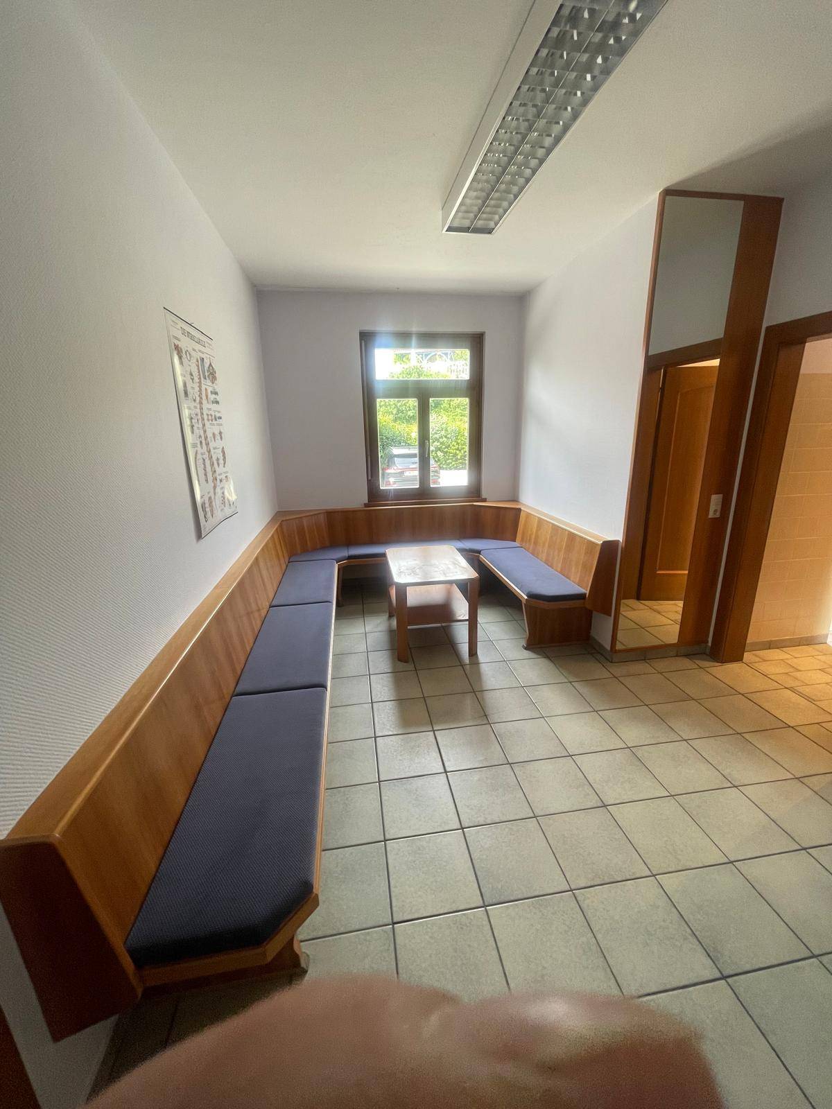 Warteraum mit U-förmiger Sitzbank, kleinem Holztisch und Fenster mit Blick ins Grüne.