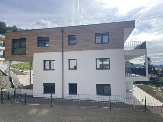 Seitenansicht eines modernen Neubaus mit Balkonen und Holzfassade unter bewölktem Himmel.