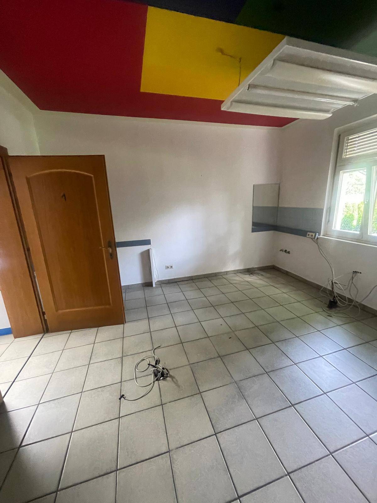 Leerer Raum mit Fliesenboden, Fenster und einer Decke mit farbigen Paneelen.
