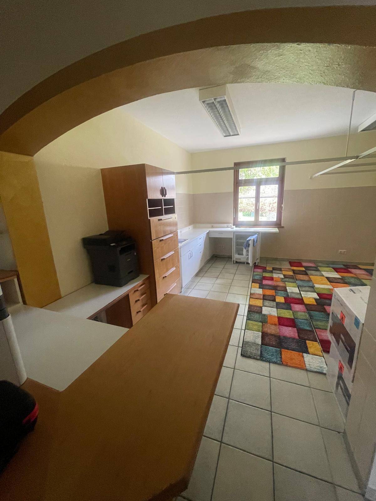 Arbeitszimmer mit Schreibtisch, Drucker, Schränken und einem bunten Teppich auf Fliesenboden.