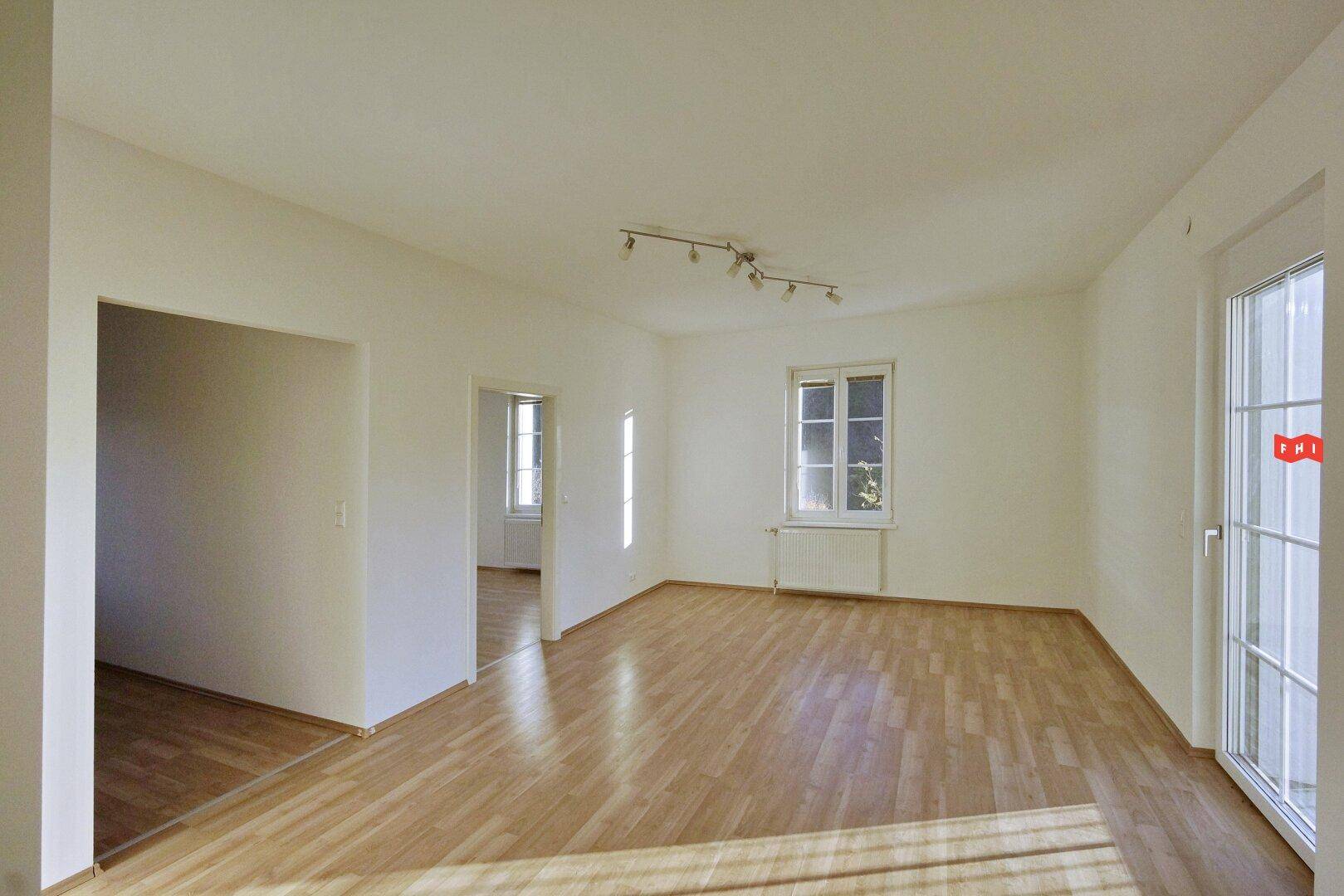 Helles Zimmer mit Holzboden, großen Fenstern und Zugang zu einem Außenbereich.