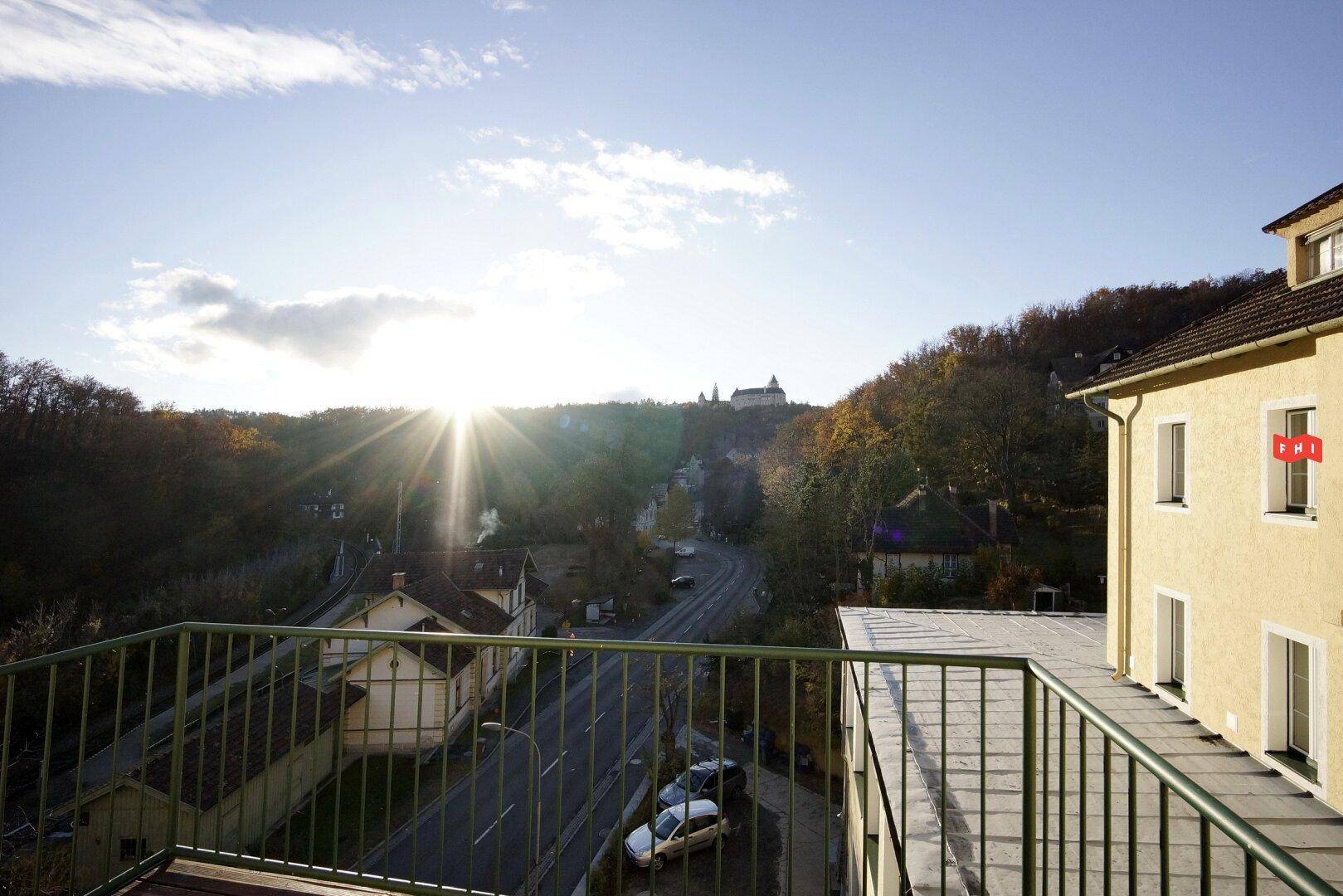Balkon mit Metallgeländer und weitem Blick über eine Stadt und bewaldete Hügel bei Sonnenuntergang.