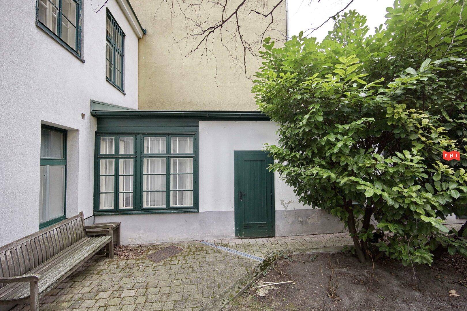 Ruhiger Innenhof mit weißer Fassade, grünen Fenstern, einer Holzbank und üppigem Grün.