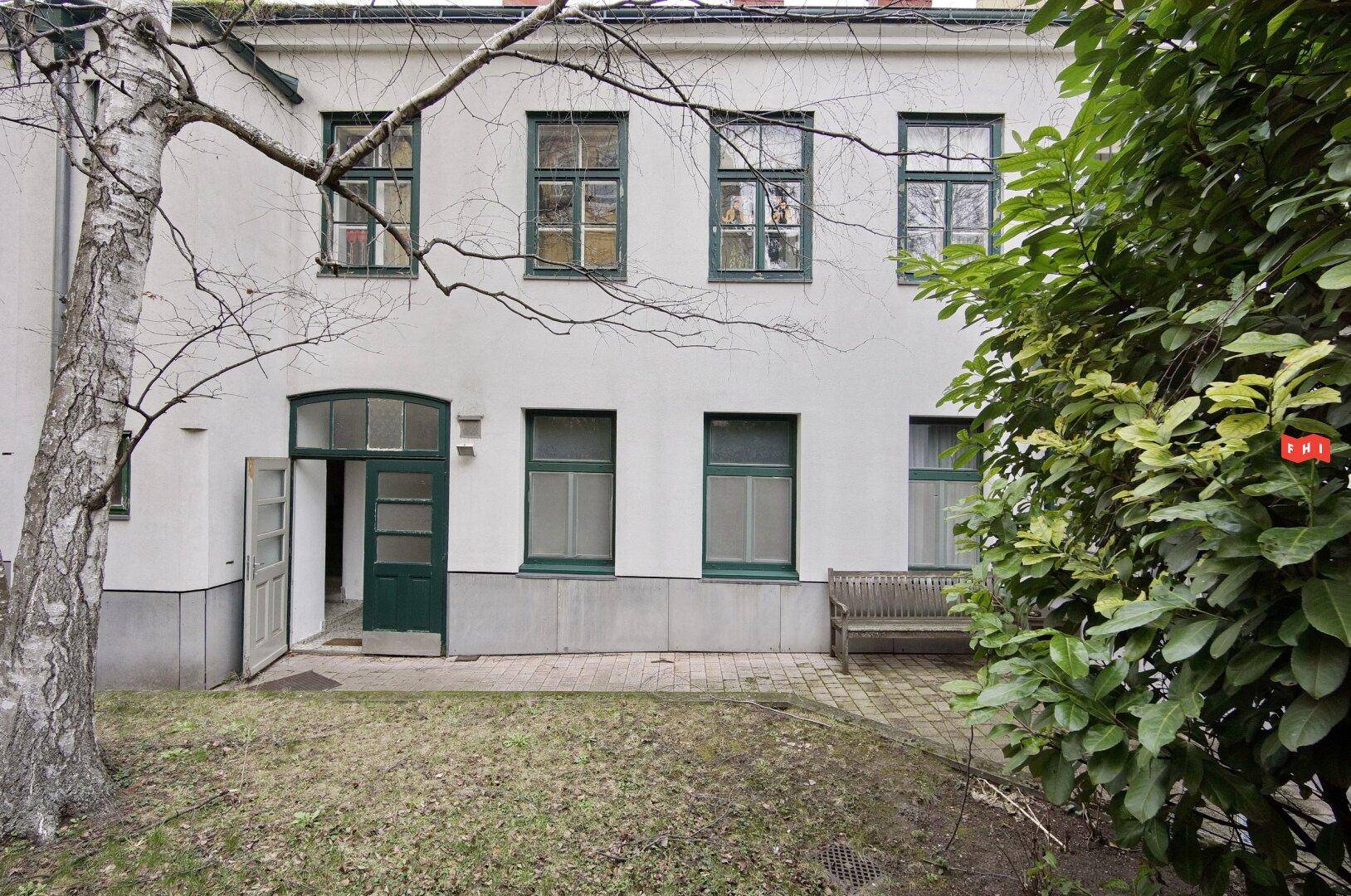 Innenhof mit weißer Fassade, grünen Fenstern, einer Holzbank und einem Baum im Vordergrund.