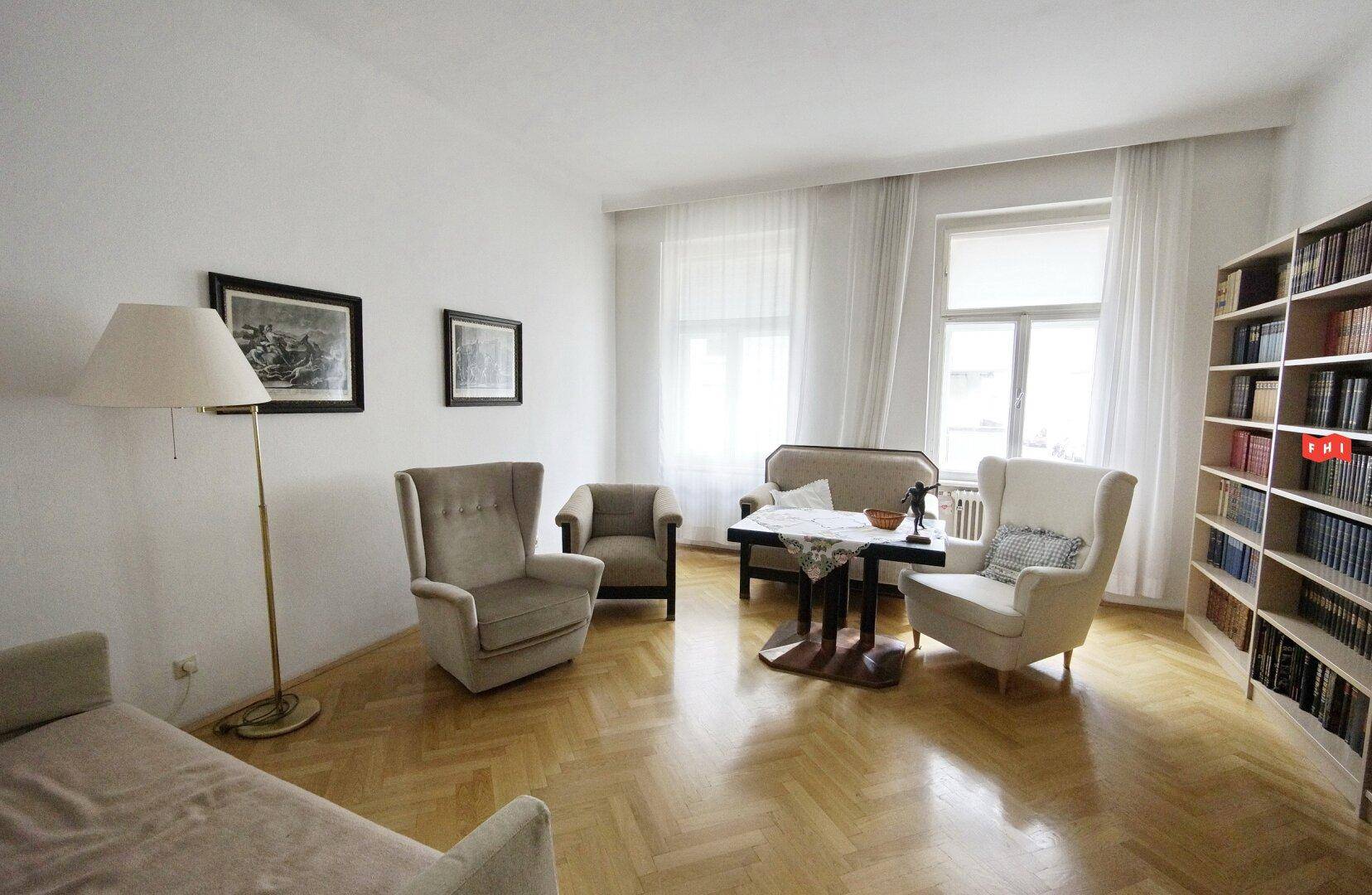 Helles Wohnzimmer mit Fischgrätparkett, bequemen Sesseln, einem Bücherregal und großen Fenstern.