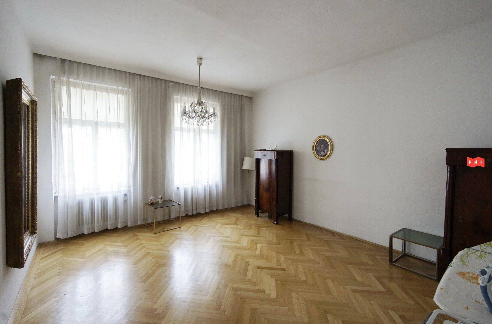 Geräumiges Zimmer mit hellem Fischgrätparkett, zwei großen Fenstern und einem eleganten Kronleuchter.