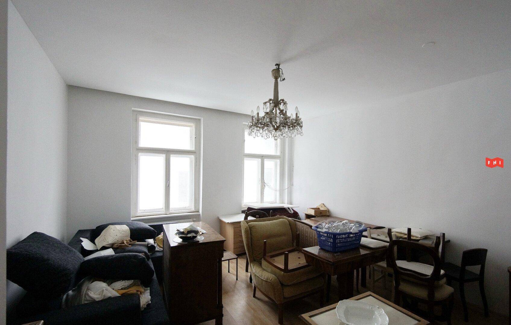 Wohnraum mit Sofa, verschiedenen Möbelstücken und zwei Fenstern, die für natürliches Licht sorgen.