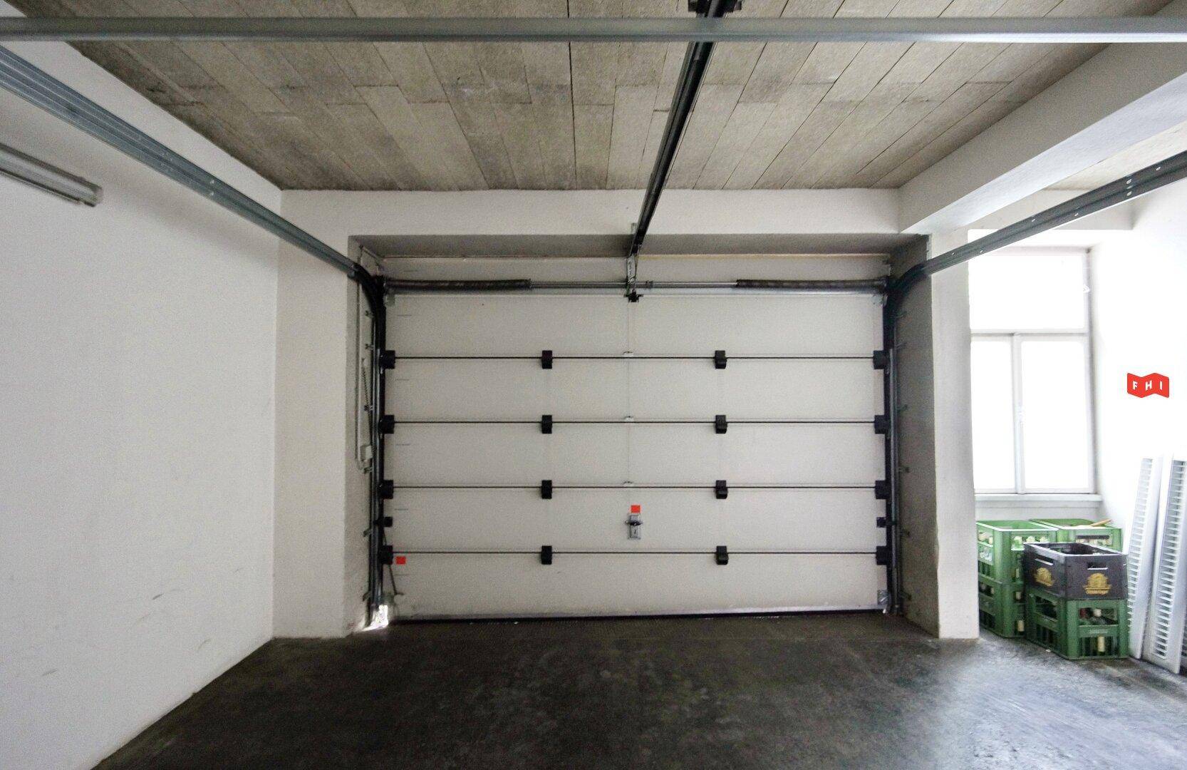 Geräumige Garage mit großem weißen Tor, Betonboden und sichtbarer Deckenkonstruktion.