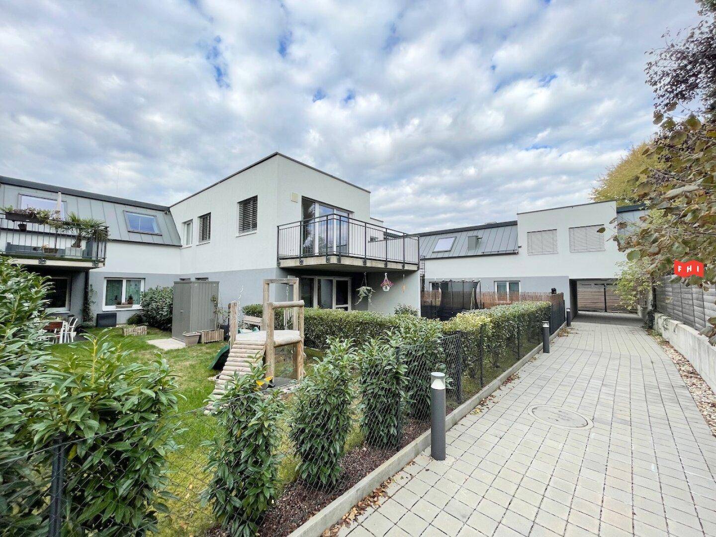 Rückansicht eines modernen Wohnkomplexes mit Gärten, Balkonen und einem kleinen Spielplatz unter bewölktem Himmel.