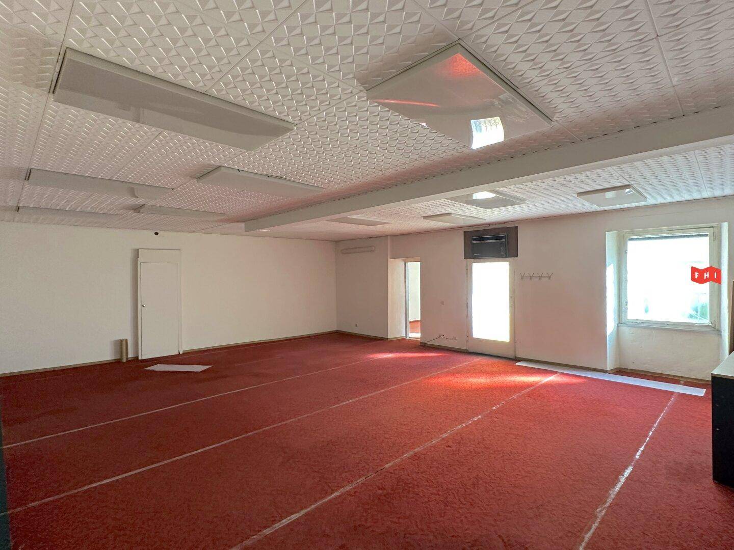 Weitläufiger Innenraum mit rotem Teppichboden, vielen Deckenleuchten und einem Fensterbereich.
