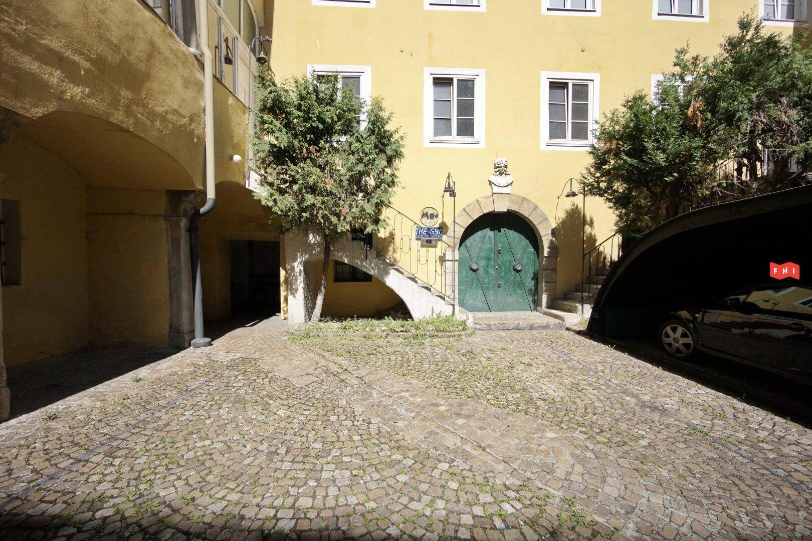 Historischer Innenhof mit Kopfsteinpflaster, gelber Fassade, grünem Tor und überdachtem Stellplatz.
