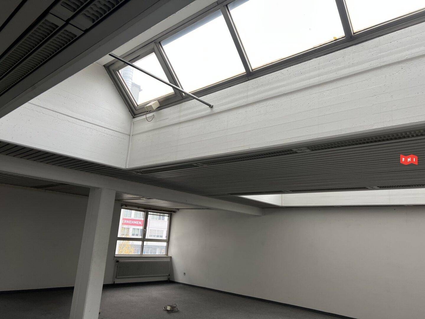 Heller Büroraum mit Teppichboden, großen Dachfenstern und einem Heizkörper unter dem Fenster.