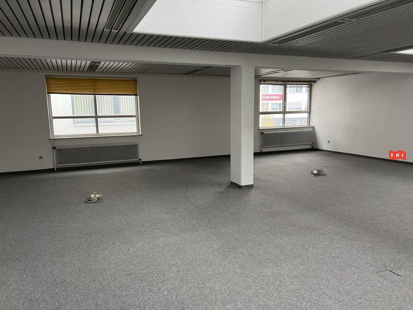 Großer Büroraum mit grauem Teppichboden, Säulen und Fenstern mit Blick auf andere Gebäude.