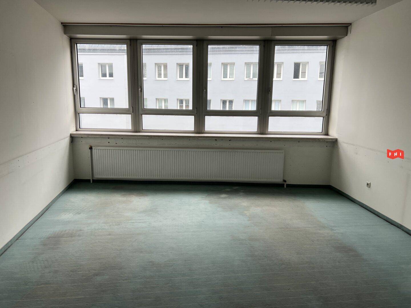 Leerer Büroraum mit blaugrünem Teppichboden, breiten Fenstern und einem Heizkörper darunter.
