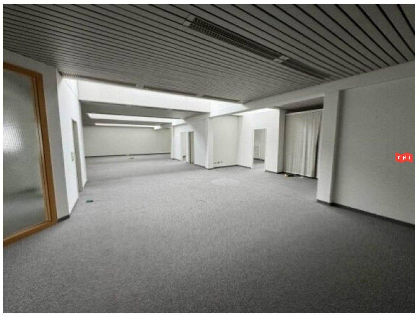 Geräumiger Bürobereich mit grauem Teppichboden, abgehängter Decke und mehreren Türen.