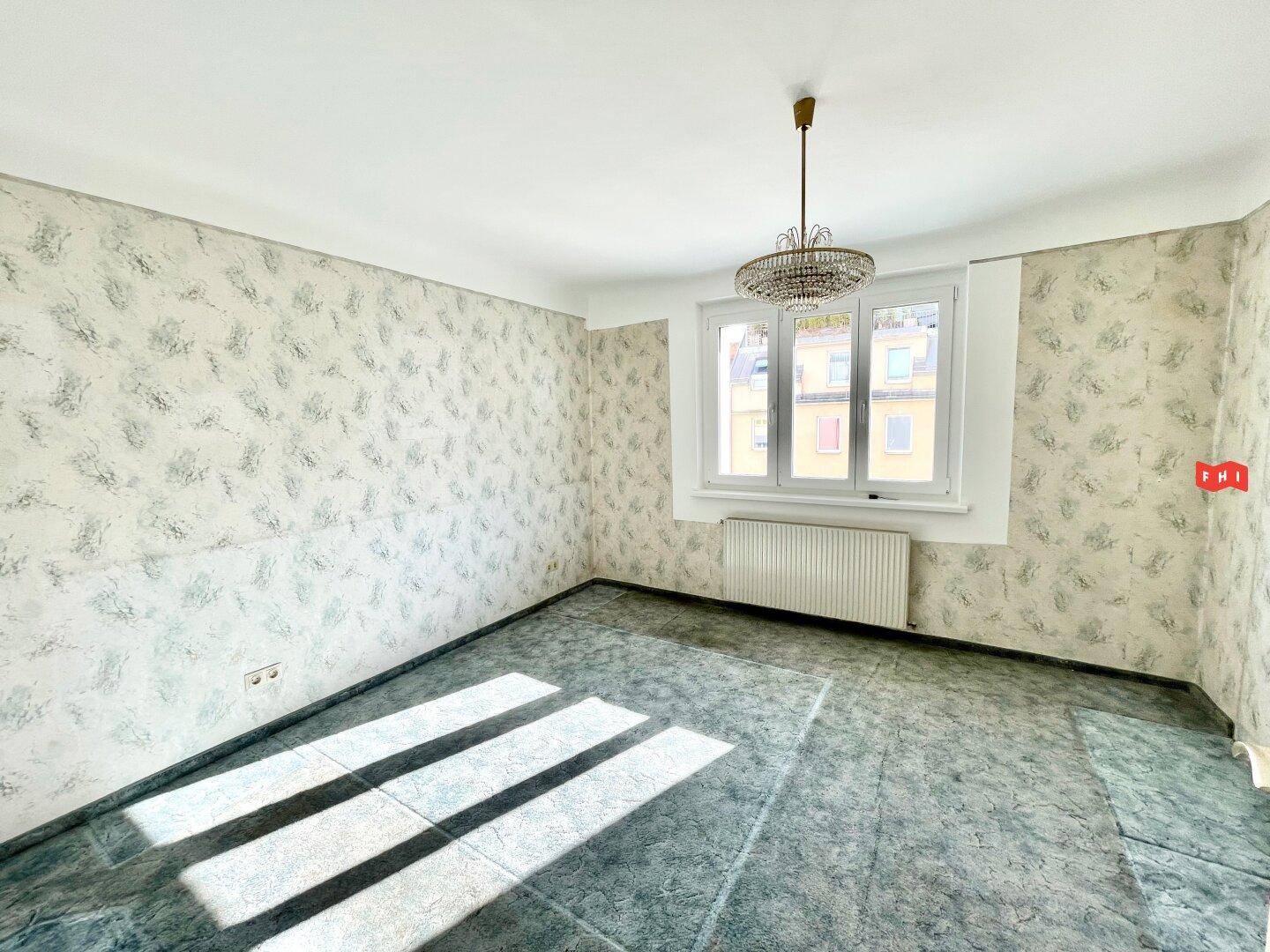 Leeres Zimmer mit gemusterter Tapete und grünem Teppichboden, durchflutet von Sonnenlicht.