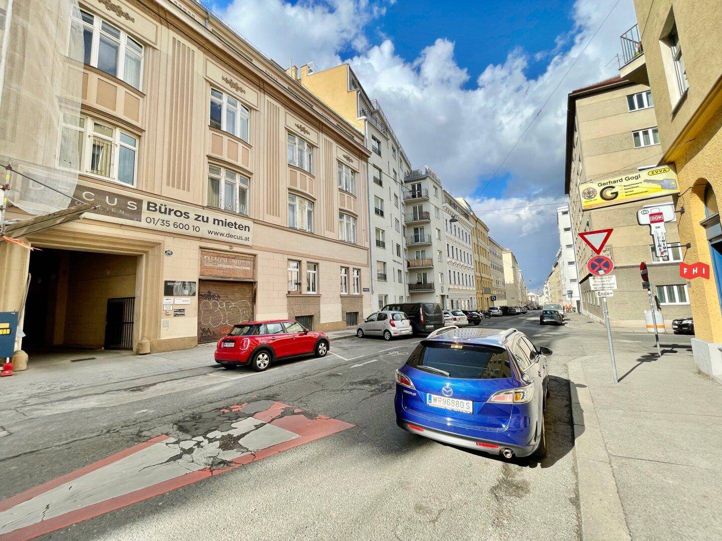 Straßenansicht mit parkenden Autos und Gebäuden unter einem bewölkten Himmel.
