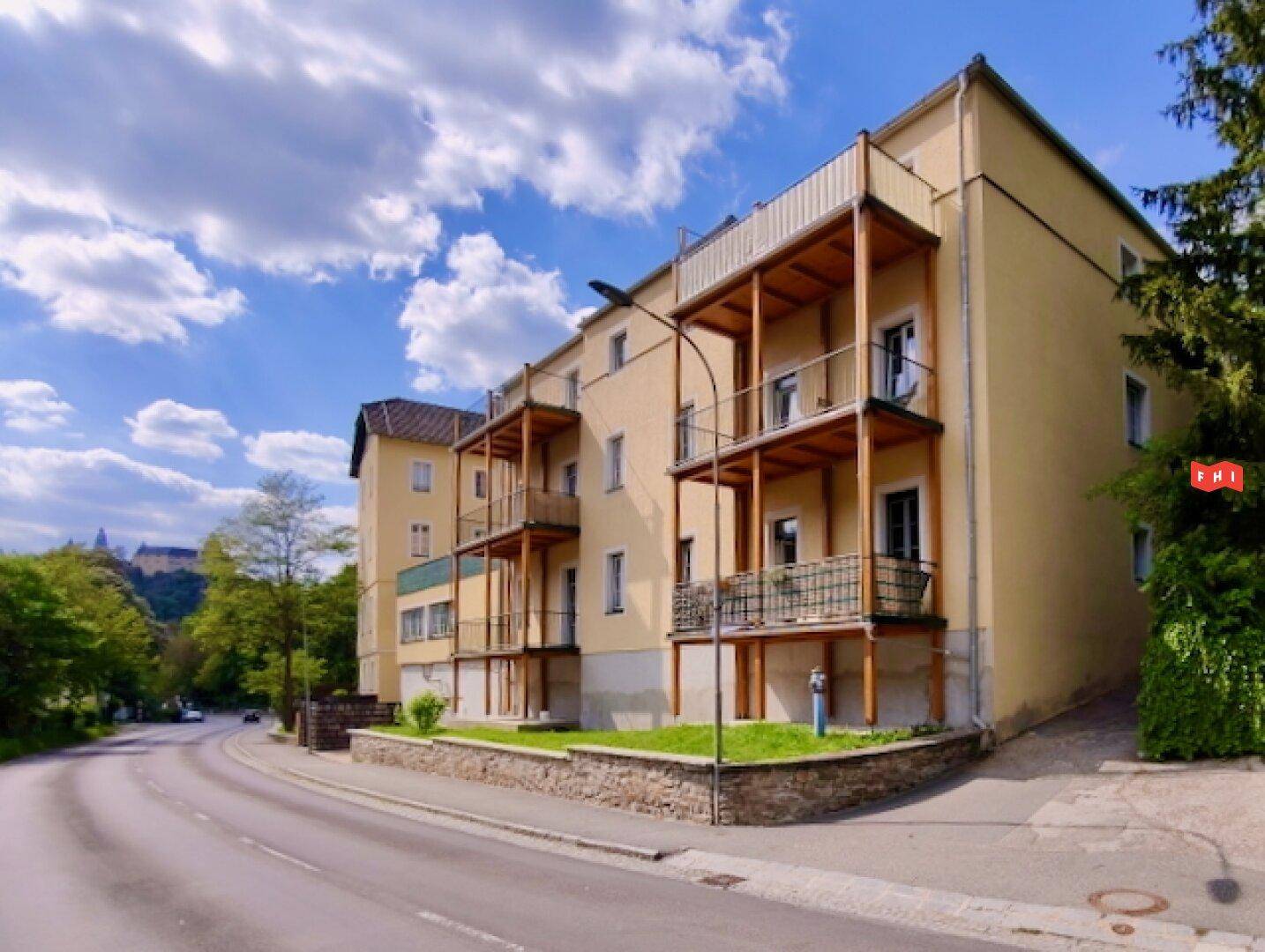 Mehrfamilienhaus mit mehreren Balkonen und gelber Fassade an einer Straße mit grüner Umgebung.