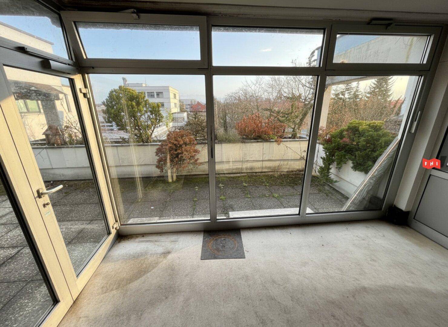 Verglaster Terrassenbereich mit Zugang zu einer Außenfläche, umgeben von Bäumen und Gebäuden.