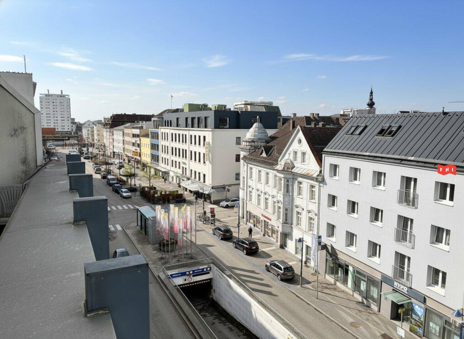 Weitläufiger Blick über eine belebte Stadtstraße mit Gebäuden, Autos und einem U-Bahn-Eingang.